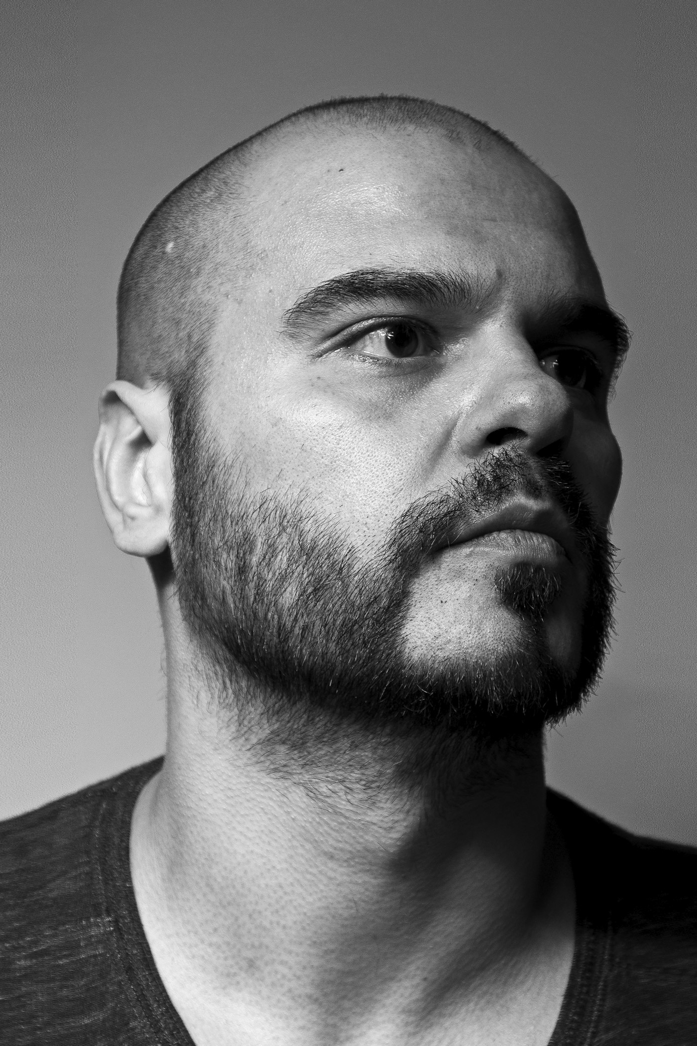 Bald man in a grey crew-neck shirt photographed in a dark editorial style.